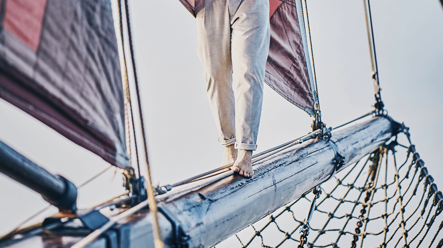 Person mit hochgekrempelten Hosen steht barfuß auf dem Bugspriet eines Segelschiffs, mit gespannten Segeln im Hintergrund.