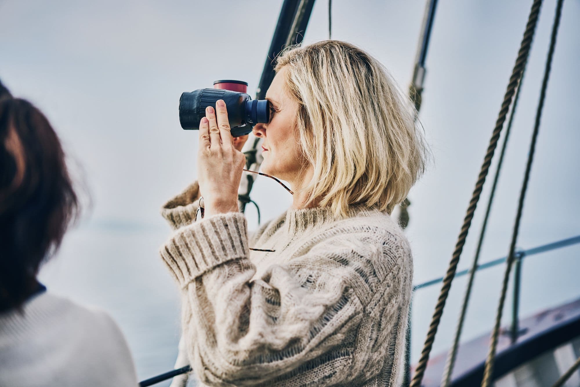 Eine Person an Bord eines Segelschiffs blickt durch ein Fernglas in die Ferne.