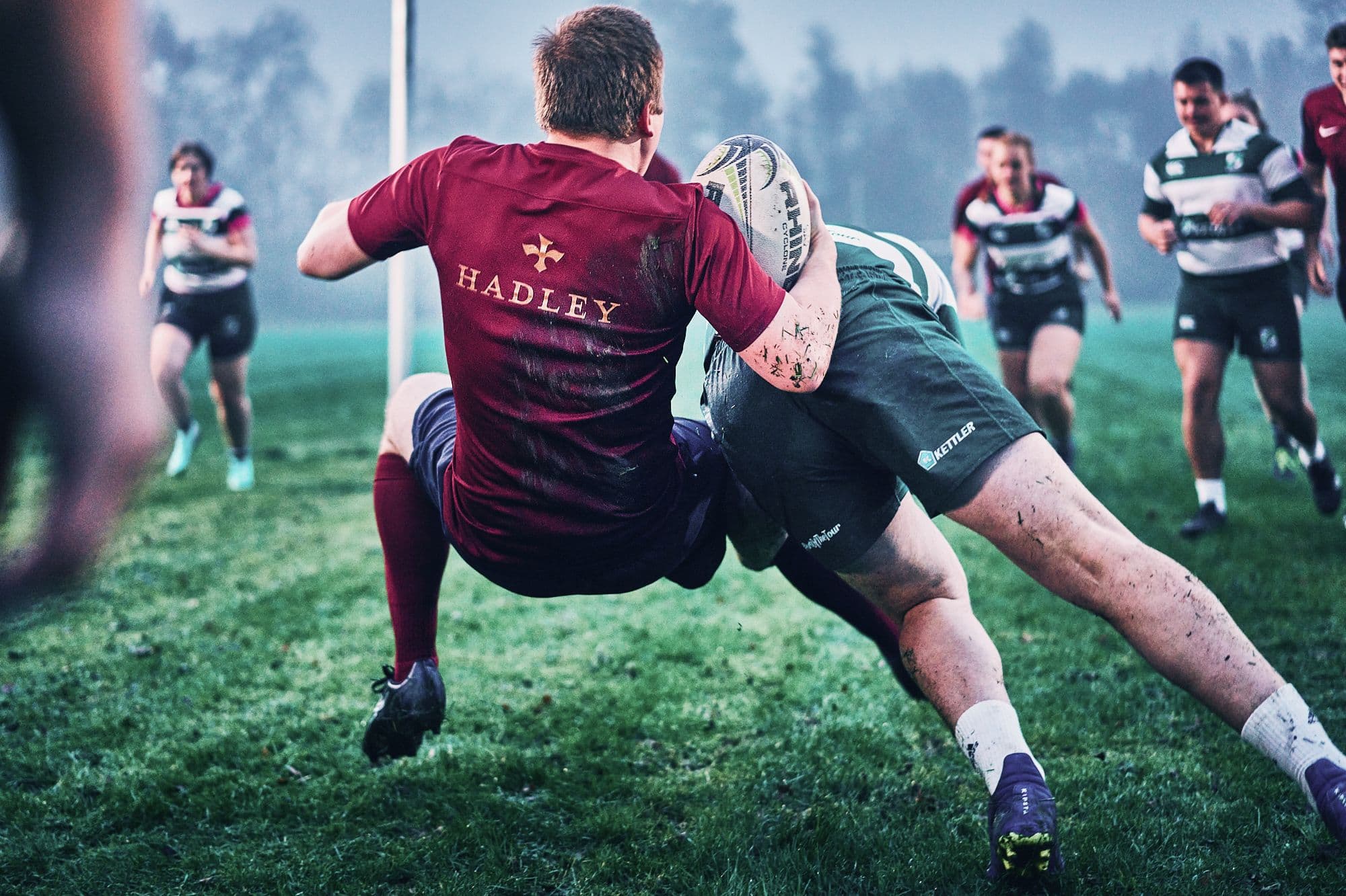 Ein HADLEY-Teamspieler in einem Rugby-Match wird beim Tackling festgehalten, während Mitspieler und Gegner im Hintergrund heranstürmen.