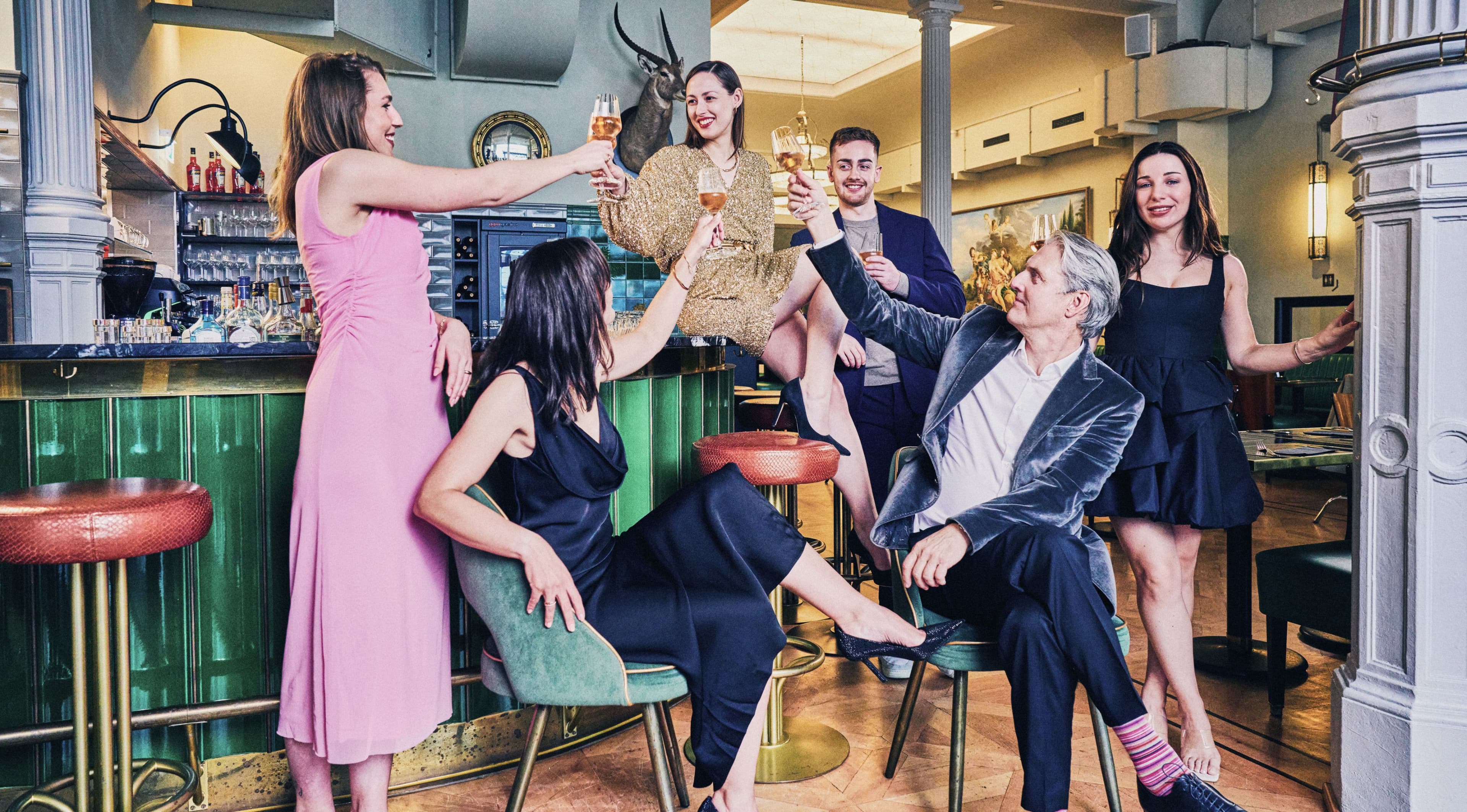 HADLEY-Teammitglieder stoßen mit Weingläsern in einer stilvollen Bar fröhlich an, elegant gekleidet und in geselliger Atmosphäre.