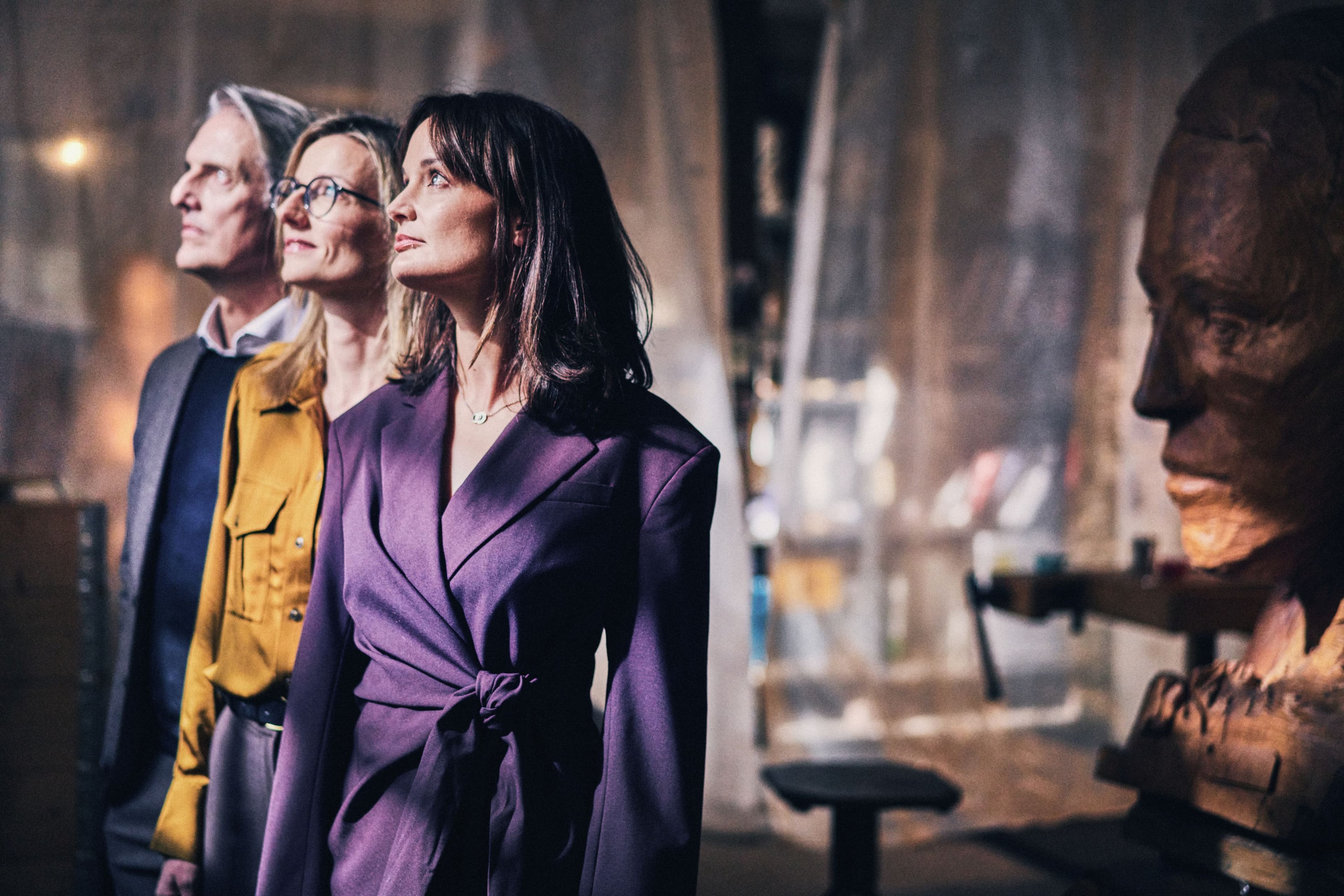 Drei Menschen in eleganter Kleidung blicken fokussiert nach vorne, im Hintergrund eine Holzskulptur in einem kunstvollen Ambiente.