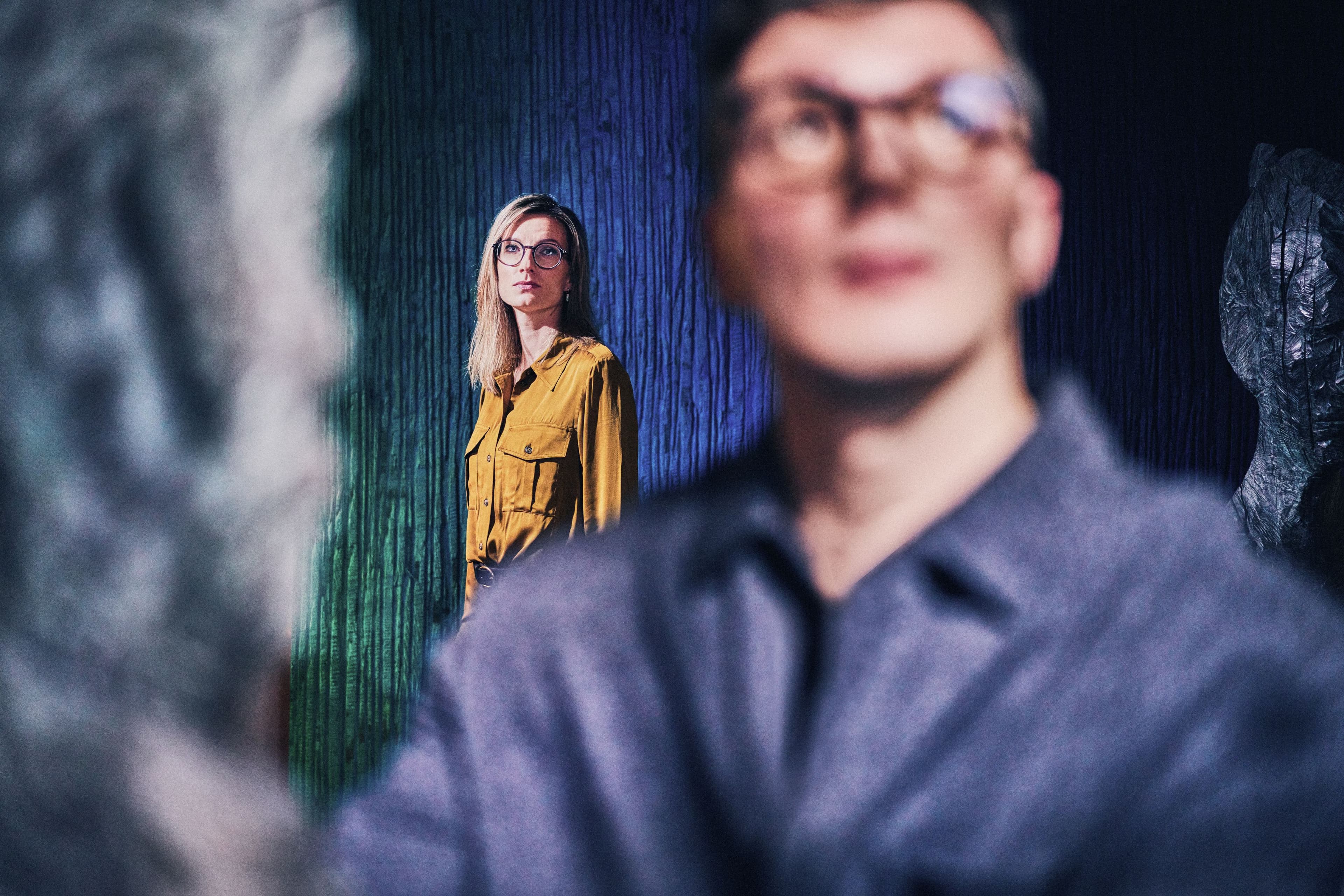 Eine Person in gelbem Hemd steht vor einem blauen Hintergrund und blickt fokussiert nach vorne, während eine unscharfe Figur im Vordergrund zu sehen ist.