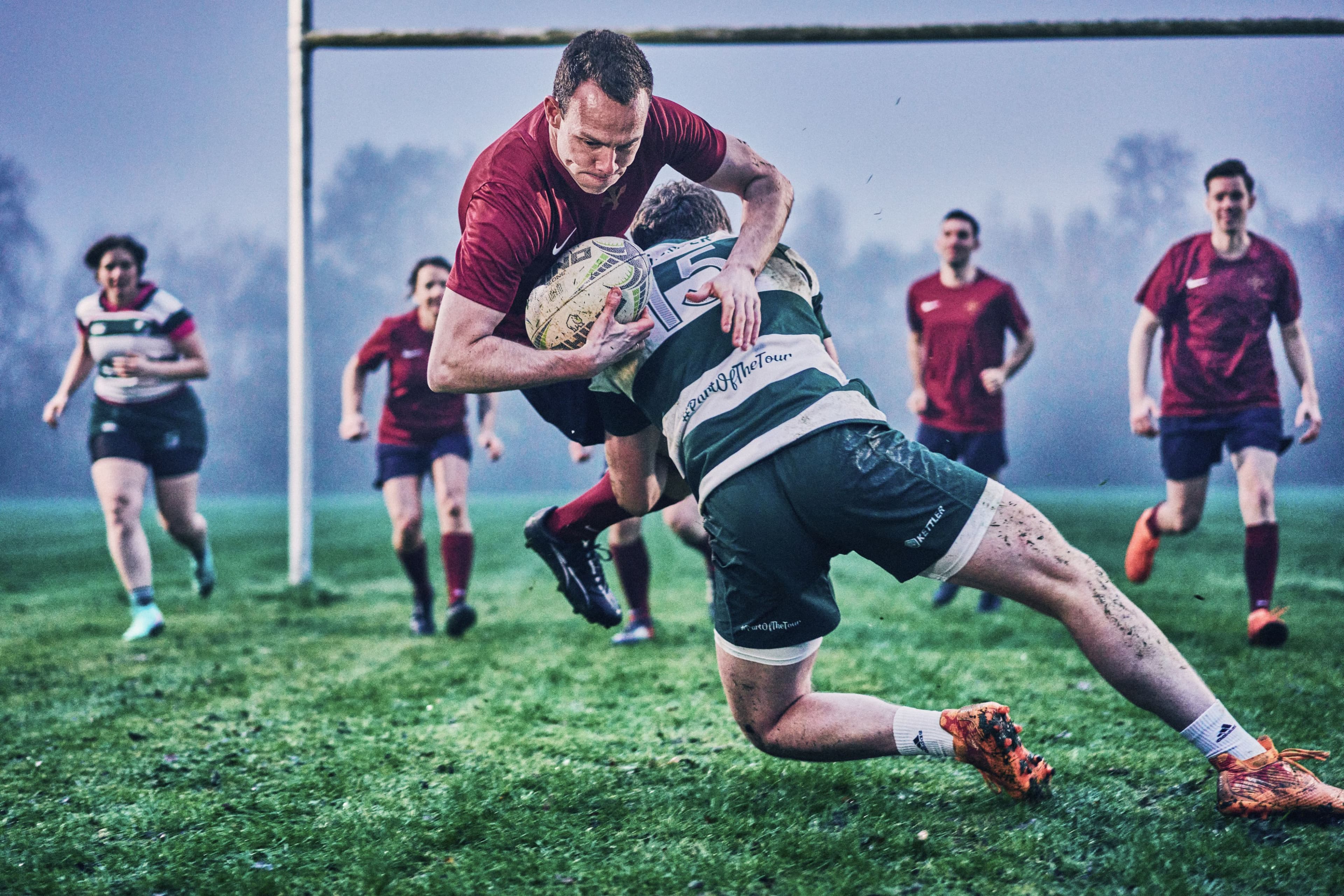 Ein Spieler des HADLEY-Teams in einem Rugby-Spiel wird von einem Gegner getackelt, während Teammitglieder im Hintergrund mitlaufen.