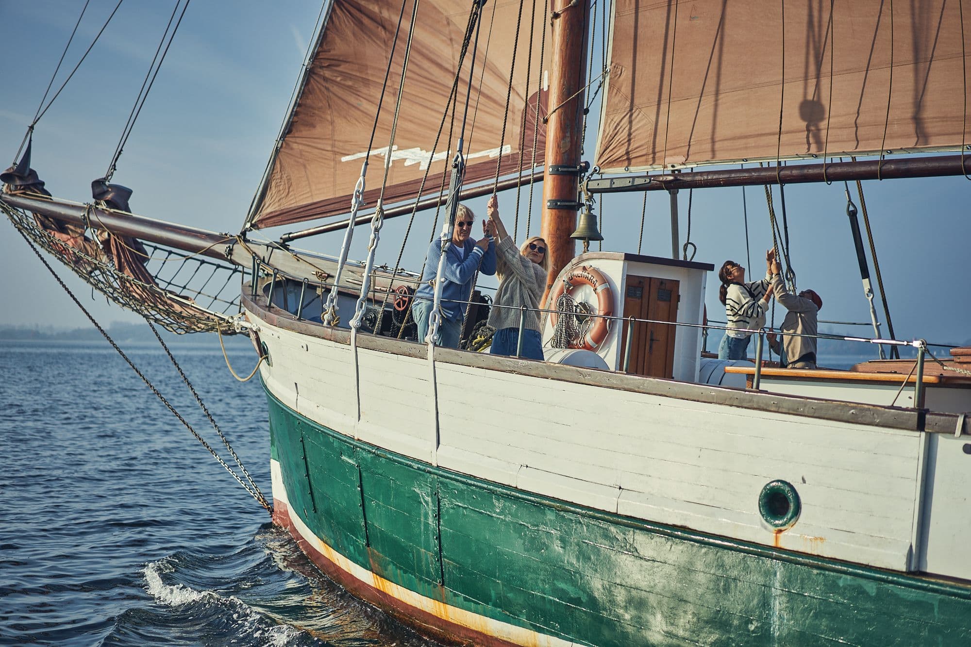 Menschen an Bord eines Segelschiffs, die gemeinsam die Segel setzen, unter klarem Himmel auf offenem Wasser. Menschen an Bord eines Segelschiffs, die gemeinsam die Segel setzen, unter klarem Himmel auf offenem Wasser.