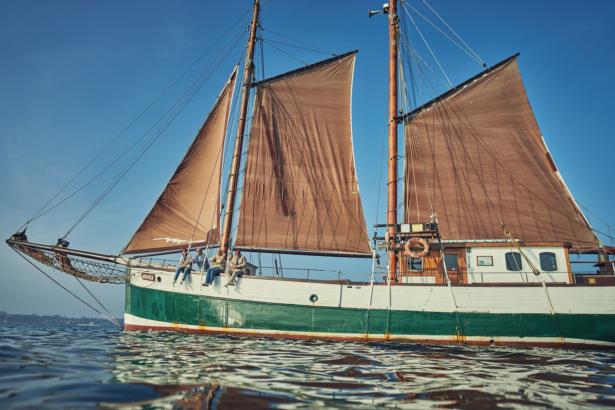 Segelschiff mit gesetzten braunen Segeln auf offenem Wasser, mit vier Menschen, die an der Reling sitzen. Segelschiff mit gesetzten braunen Segeln auf offenem Wasser, mit vier Menschen, die an der Reling sitzen.